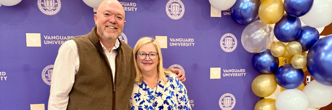 Man and woman stand in front of a step and repeat banner for Vanguard University and smile, celebrating David's five year anniversary at Vanguard University